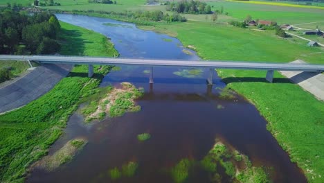 aerial view of a venta river on a sunny summer day, lush green trees and meadows, beautiful rural landscape, wide angle drone shot moving forward over the white concrete bridge, car passes by