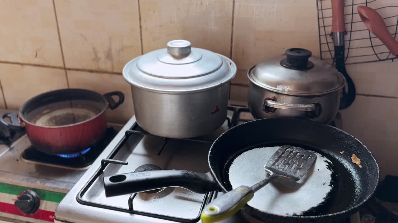 Close view of aluminum pots and nonstick frying pan on a lit gas stove, spatula resting in pan, tiled backsplash and utensils, everyday home kitchen cooking setup