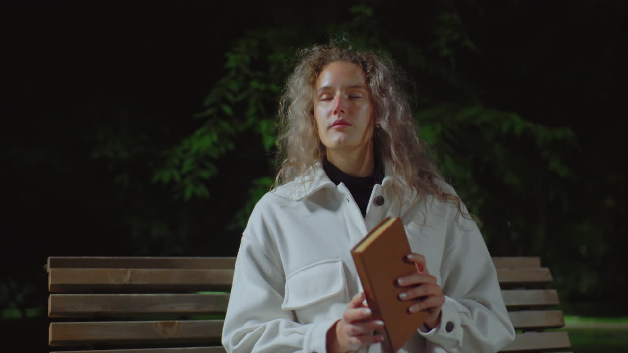 Young girl wearing white coat seated outside during cool evening, taking deep breath while preparing to open book to read, surrounded by calm dark background and faint greenery