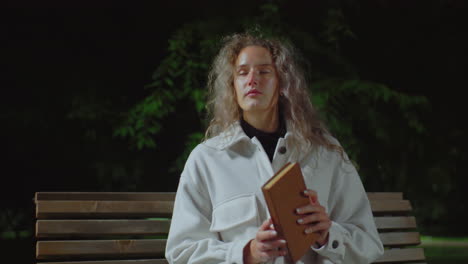 young girl wearing white coat seated outside during cool evening, taking deep breath while preparing to open book to read, surrounded by calm dark background and faint greenery