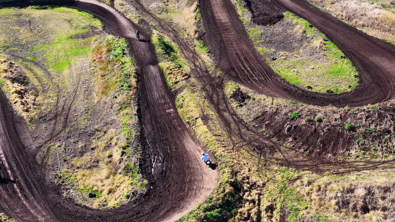A dirt bike rider accelerates and jumps along a winding motocross track in a rural, sunlit Australian bush setting, captured by aerial drone