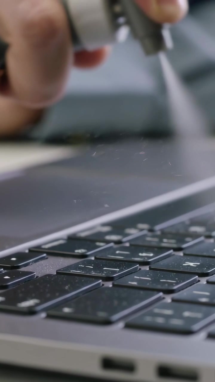 Vertical video: Spraying hand pressing spray nozzle cleaning laptop keyboard at desk, mist visible