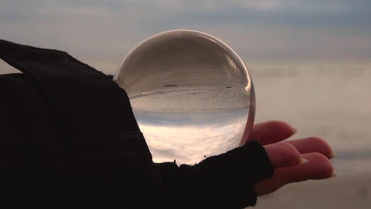 mano femminile con guanto senza dita che tiene una sfera di cristallo che riflette capovolta una spiaggia e un paesaggio marino in una giornata invernale nuvolosa e ventosa