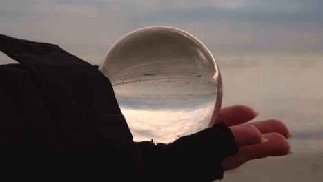 female hand with fingerless glove holding a crystal ball reflecting upside down a beach and sea landscape on a cloudy and windy day of winter