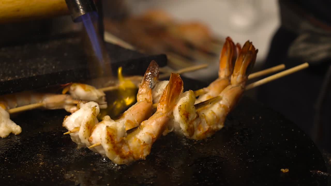 Close-up of shrimp skewers being flame-torched on a grill plate by a gloved hand.