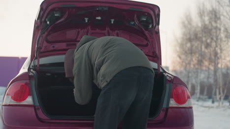 back view of student in winter outfit accessing car boot while parked near trees with residential building visible in distant background, focused posture and casual vehicle interaction on cold day