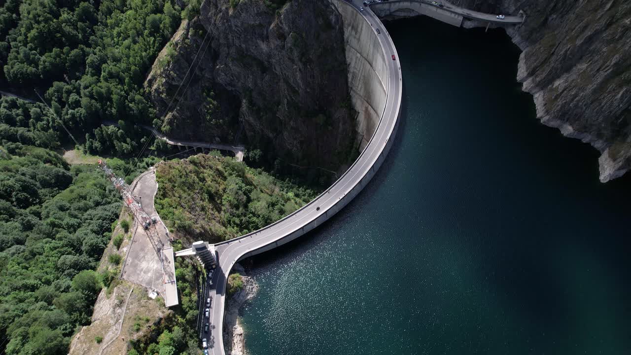 Aerial shot of Vidraru Dam in Romania, captured at an angle while circling left. Blue lake, dramatic curves, and green mountain backdrop in 4K.
