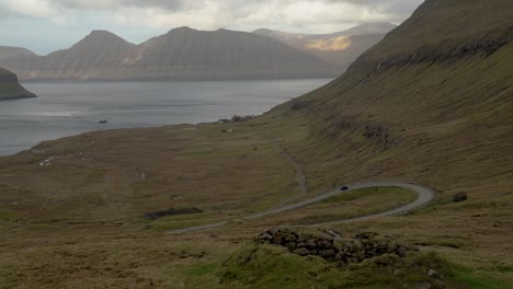 Car-driving-on-mountain-road-near-Hellurnar-town-with-Oyndarfjordur-Fjord-on-Eysturoy-Island---Kalsoy-island-in-background