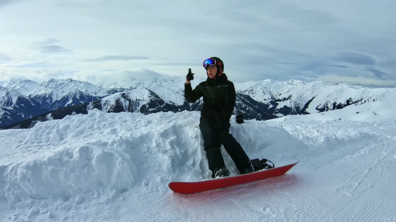 snowboarder adolescente tomando selfie sentado en una nieve rodeada de montañas