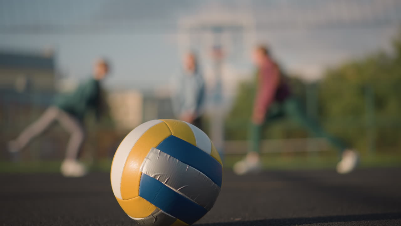 한 배경으로 바닥에 놓인 배구의 클로즈업으로 사람들이 활기찬 운동 활동과 함께 야외 스포츠 경기장에서 운동하고 스트레칭하는 것을 보여줍니다.