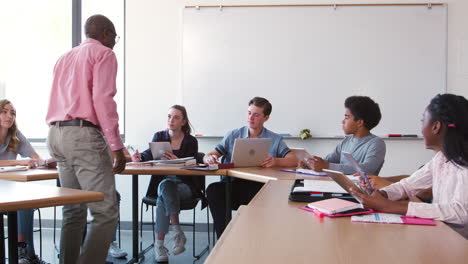 Profesor-De-Secundaria-Hablando-Con-Alumnos-Usando-Dispositivos-Digitales-En-Clase-De-Tecnología