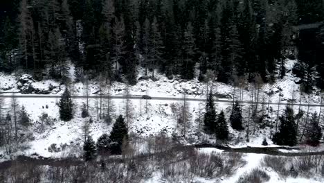 Aerial-side-view-of-a-small-car-driving-on-a-snow-covered-mountain-road-surrounded-by-forests-during-a-cloudy-winter-day-with-snowfall-in-Switzerland