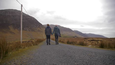 pareja se toma de la mano mientras camina por la carretera en connemara, vista trasera de ángulo bajo