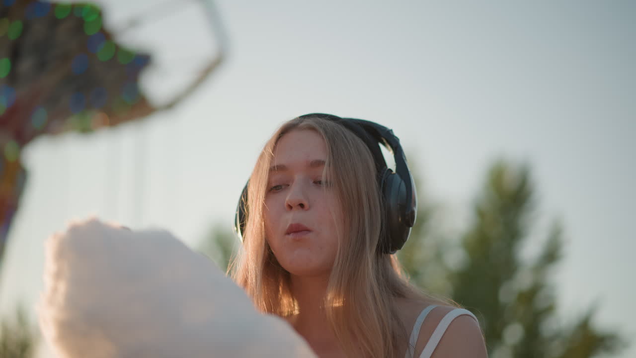 Adolescente sognante in stile carnevalesco, con cuffie, che guarda il tramonto, addentando lentamente dello zucchero filato, sorriso bizzarro, posa da influencer, aspirante cantante, primo piano emozionante, luce solare soffusa, serata nostalgica