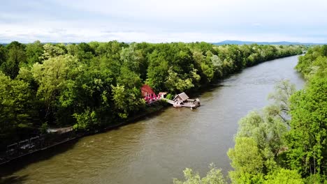 Luftaufnahme-Einer-Schiffsmühle-über-Dem-Fluss-Mura,-Wälder-Auf-Beiden-Seiten,-Bewölkter-Himmel,-Österreich,-Slowenien,-Europa