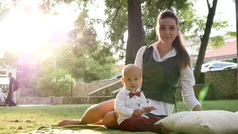 young mother sits with cute baby in suit and bow tie on green lawn in park