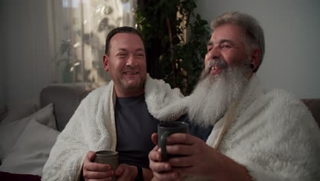 a happy elderly man with gray hair and a lush gray beard is wrapped in a white woolen blanket and drinks hot tea with his brunette boyfriend in a gray gray t-shirt on the sofa in the evening in a modern apartment