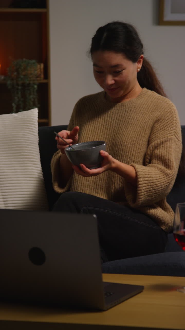 Video verticale di una donna a casa seduta sul divano che mangia un pasto sano vegetariano o vegano di insalata di tabbouleh con un bicchiere di vino mentre guarda uno spettacolo sul laptop