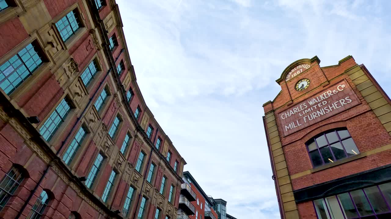 Low-angle camera pans upward along a curved red-brick building with large windows under daylight, revealing architectural details at a Leeds street junction