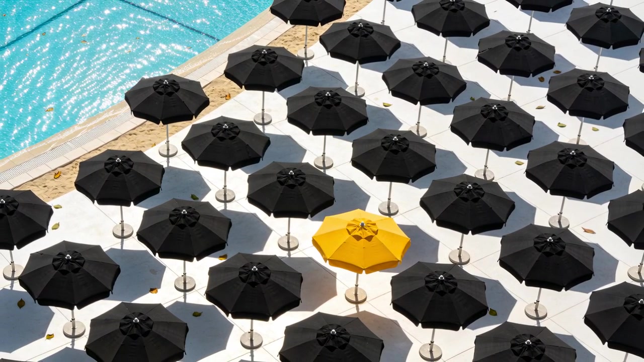 La telecamera zooma su un solarium, rivelando una griglia di ombrelloni con un ombrellone giallo, evidenziando il contrasto.