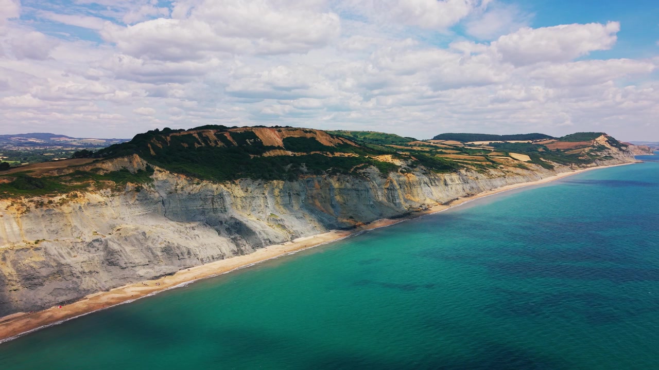 Ampie riprese aeree con drone che seguono le scogliere spettacolari e le acque turchesi della Costa Giurassica vicino a West Bay, Dorset, Inghilterra. Paesaggio costiero panoramico caratterizzato da geologia stratificata, una spiaggia tranquilla