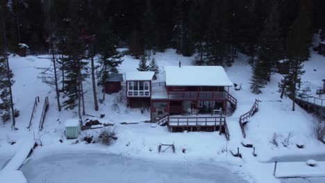 this beautiful wooden lakehouse covered in snow sits next to a frozen lake and a dense forest in british columbia