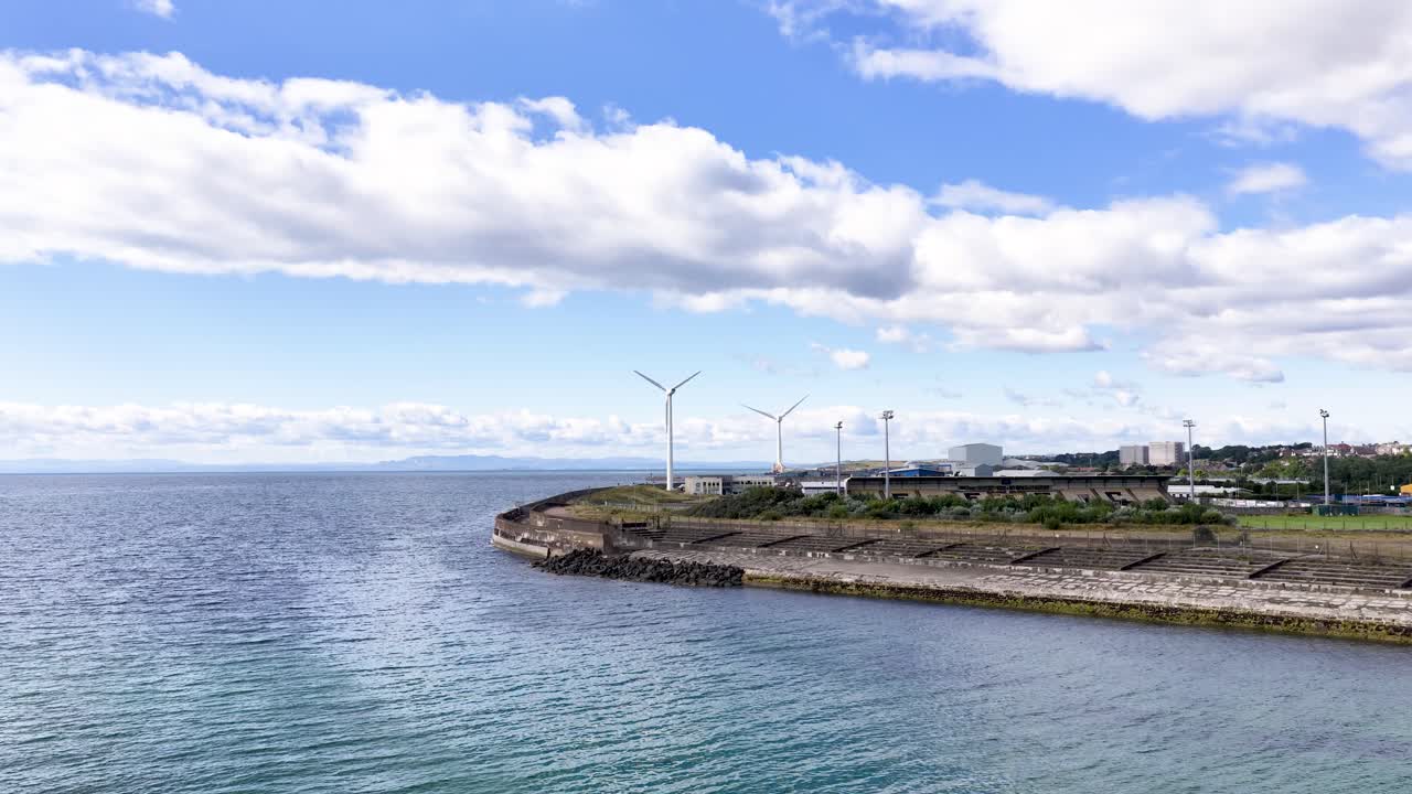 Drone pans across coastal wind turbines, blue sky, and calm sea in bright daylight