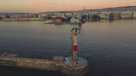 Leuchtturm-Bei-Sonnenuntergang-An-Der-Uferpromenade-Von-Thessaloniki:-Eine-Drohne-Zeigt-Einen-Leuchtturm-Mit-Vögeln,-Die-Skyline-Der-Stadt-Im-Hintergrund-–-Faszinierende-Ausblicke-Auf-Die-Schönheit-Der-Griechischen-Stadtküste.-Θεσσαλονίκη,-Ελλάδ?