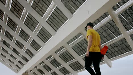 Young-Attractive-Trendy-Man-walking-with-a-skateboard-under-a-solar-panel-on-a-morning-sunny-day-with-an-urban-city-background-in-slow-motion