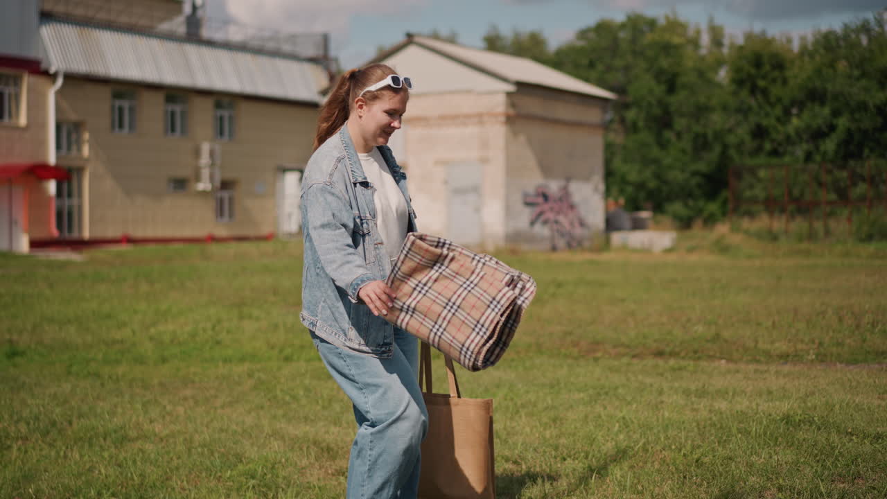 Donna che passeggia davanti a case suburbane. Acquirente che fa piccole commissioni con una borsa tote e una borsa a fantasia, giacca di jeans e jeans, fascia per capelli, pomeriggio soleggiato, cortile erboso e quartiere tranquillo, passo tranquillo