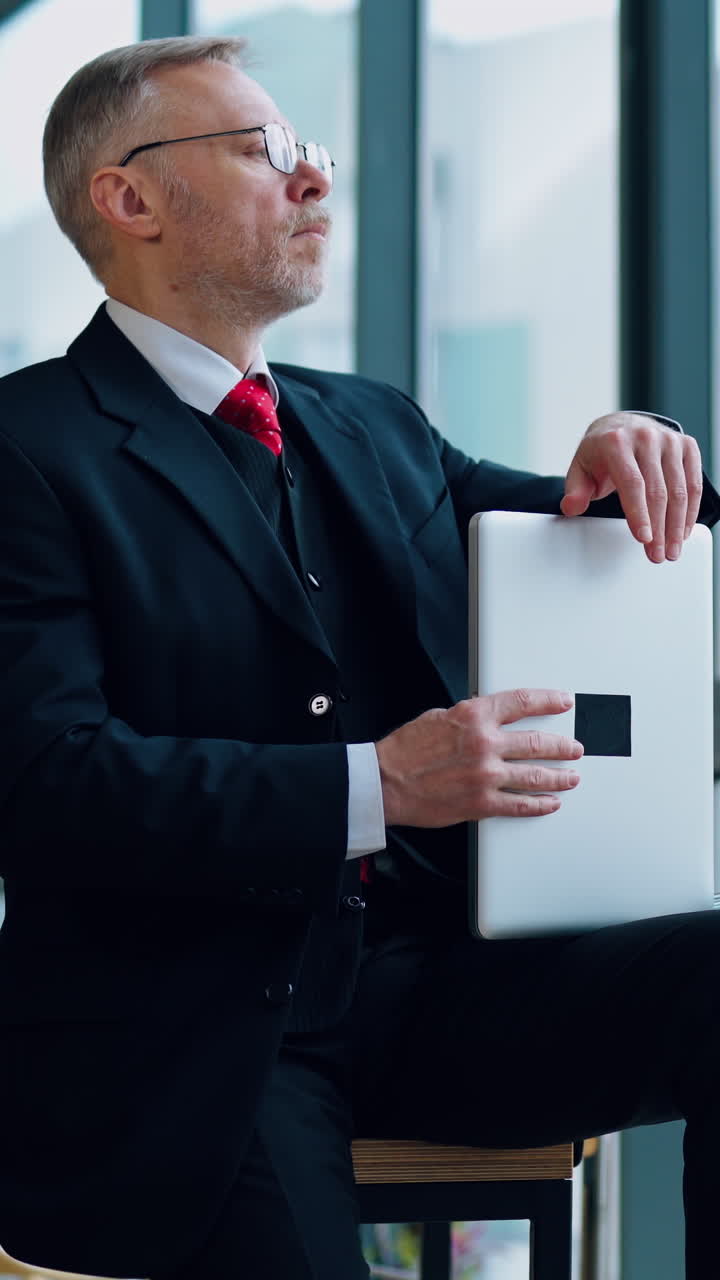 Serious entrepreneur in suit in office. Mature businessman sitting on the table with a laptop and looking through the window thoughtfully. Vertical video
