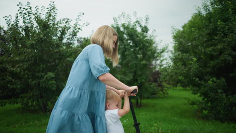 a mother rides a scooter with her young son on a walkway, using her leg to move it forward, her dress flutters gently in the wind