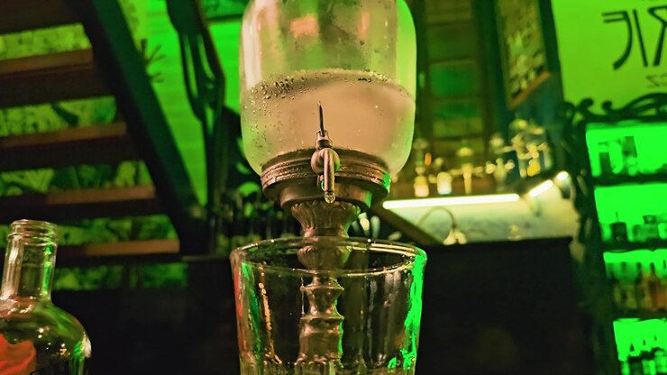 Vista dettagliata di una fontana per louche di assenzio sopra un bicchiere da cocktail, con acqua ghiacciata, beccuccio metallico vintage, illuminazione verde del bar, riflessi di bottiglie e interni scuri di un lounge di notte.