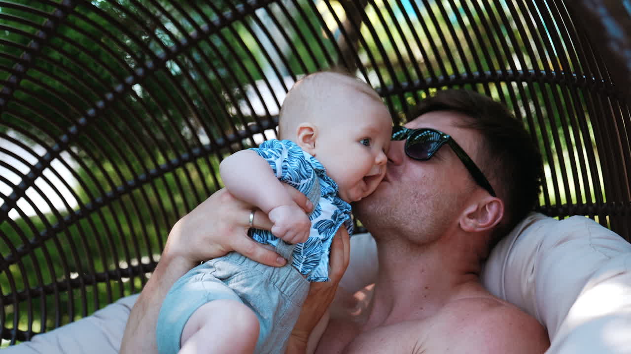Dad with his adorable little infant son. Loving father kisses his dear child sitting in the egg chair outdoors.