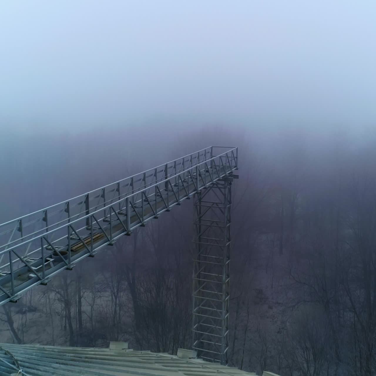 Sloping roof of an elevator tank with massive metal support over it. Drone footage along the metal pillar on the cistern. Foggy winter forest backdrop