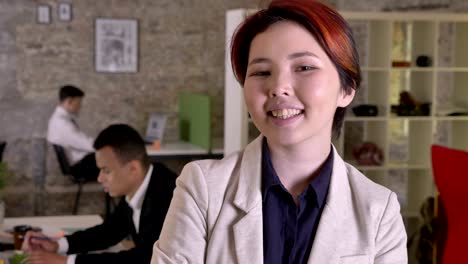 young business asian woman looking in camera in modern office, smiling, black and caucasian mans working in back