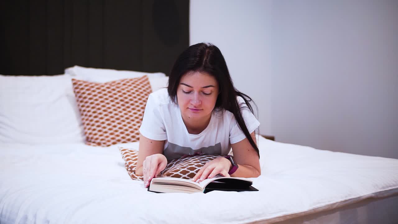 한 젊은 여성이 침실에서 검정색 양장본이 있는 책을 읽고 침대에 누워 책을 읽으며 자유 시간을 보내고 집에서 휴식을 취하고 페이지를 넘기고 페이지를 넘기고 클로즈업 샷을 찍습니다.