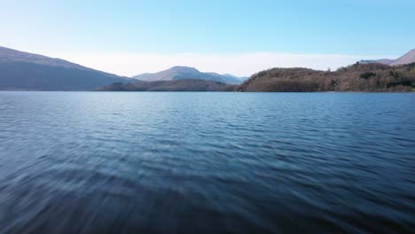 low fly along water at loch lomond and lift up pan right to ben lomond and hills on a sunny day