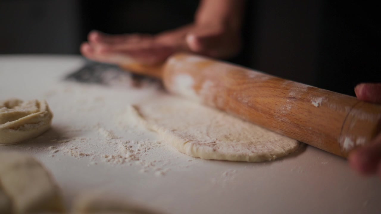Hands roll dough with wooden roller on floured surface. Focus shows texture and motion. Dusty background adds warm, homey feel. Ideal for cooking, baking, or food tutorials