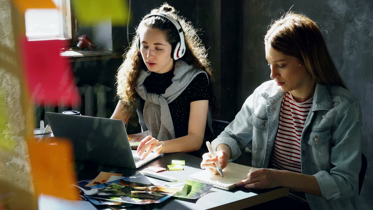un equipo de diseñadores creativos está trabajando juntos en una oficina ligera. una mujer de cabello oscuro está escuchando música y trabajando con una computadora portátil, y una mujer rubia está dibujando imágenes en el bloc de notas