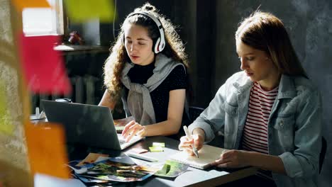 un equipo de diseñadores creativos está trabajando juntos en una oficina ligera. una mujer de cabello oscuro está escuchando música y trabajando con una computadora portátil, y una mujer rubia está dibujando imágenes en el bloc de notas
