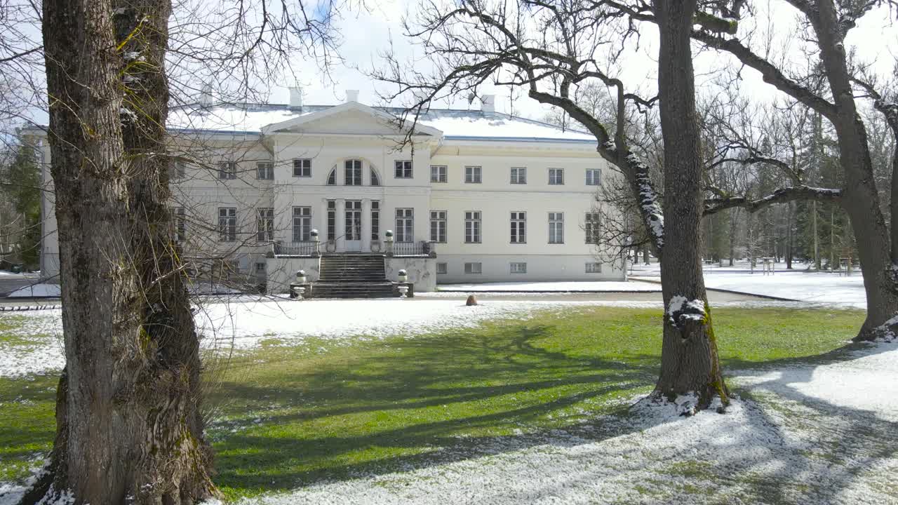 Gorgeous left to right sideways movement footage of Saku mansion or manor during a winter sunny day while white snow and ice is visible on the green grass around and on the building. White building.