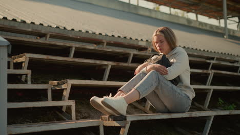 thoughtful young woman seated alone on empty stadium bleachers, hugging her bag with a reflective expression, sunlight illuminates her as she sits in contemplation