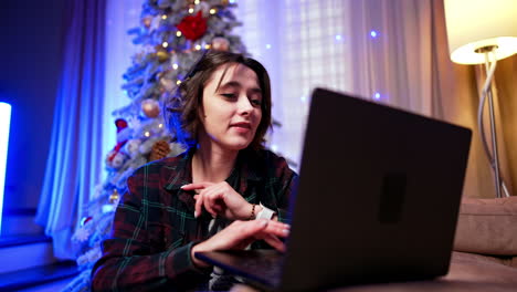 woman with laptop by tree. a young woman sits at a laptop, focused on her work, with a decorated christmas tree in the background