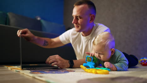 father and son at home. little baby tries to bite his toy. man opens his laptop and starts to type.