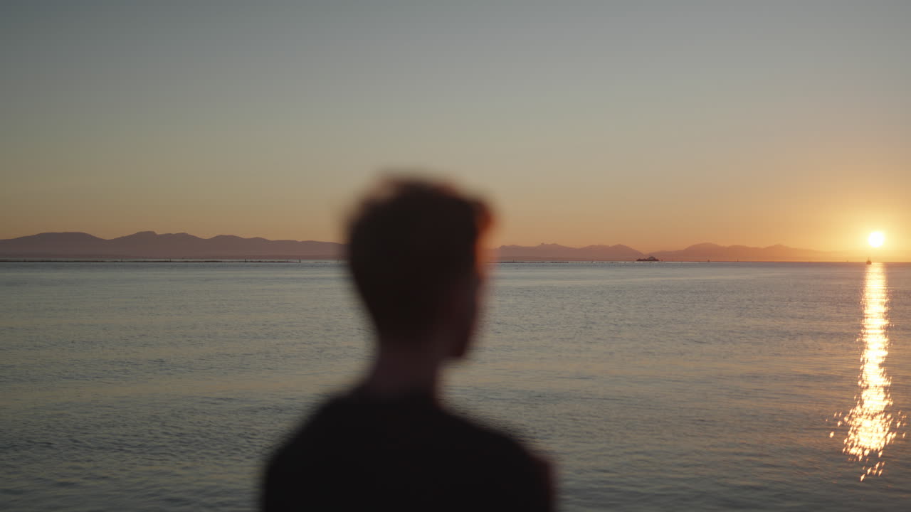 전경에 있는 초점을 잃은 사람은 먼 산이 있는 항구에서 일몰을 보고 있습니다.