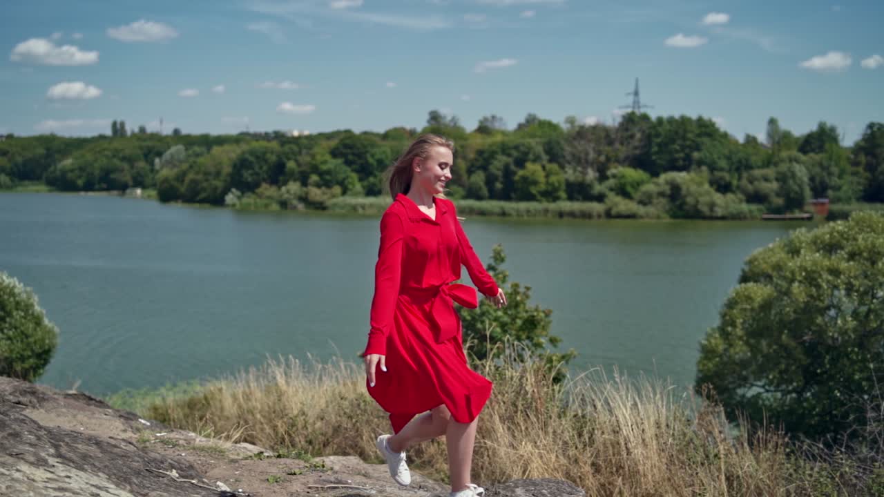 Happy woman enjoying nature in summer. Beautiful girl in red dress running and jumping merrily on the blue river background. Summer vacation.