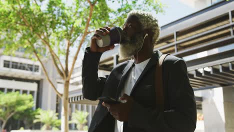 african american senior man drinking coffee and using smartphone in corporate park