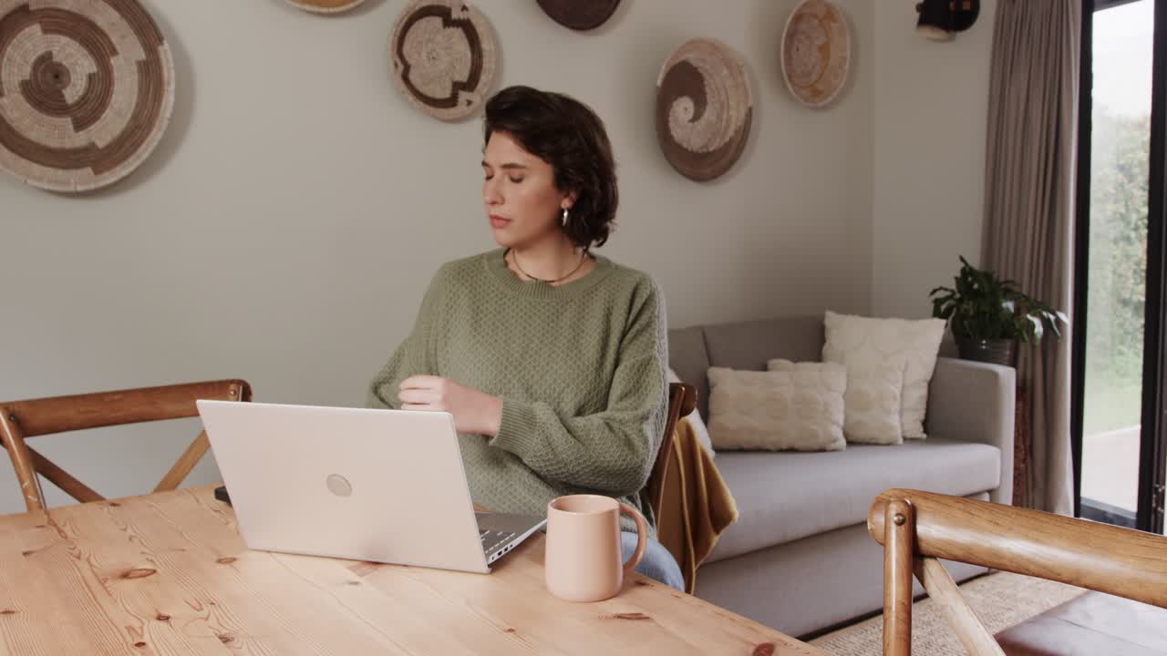 Woman in green sweater reading confusing phone message at desk before typing reply on laptop