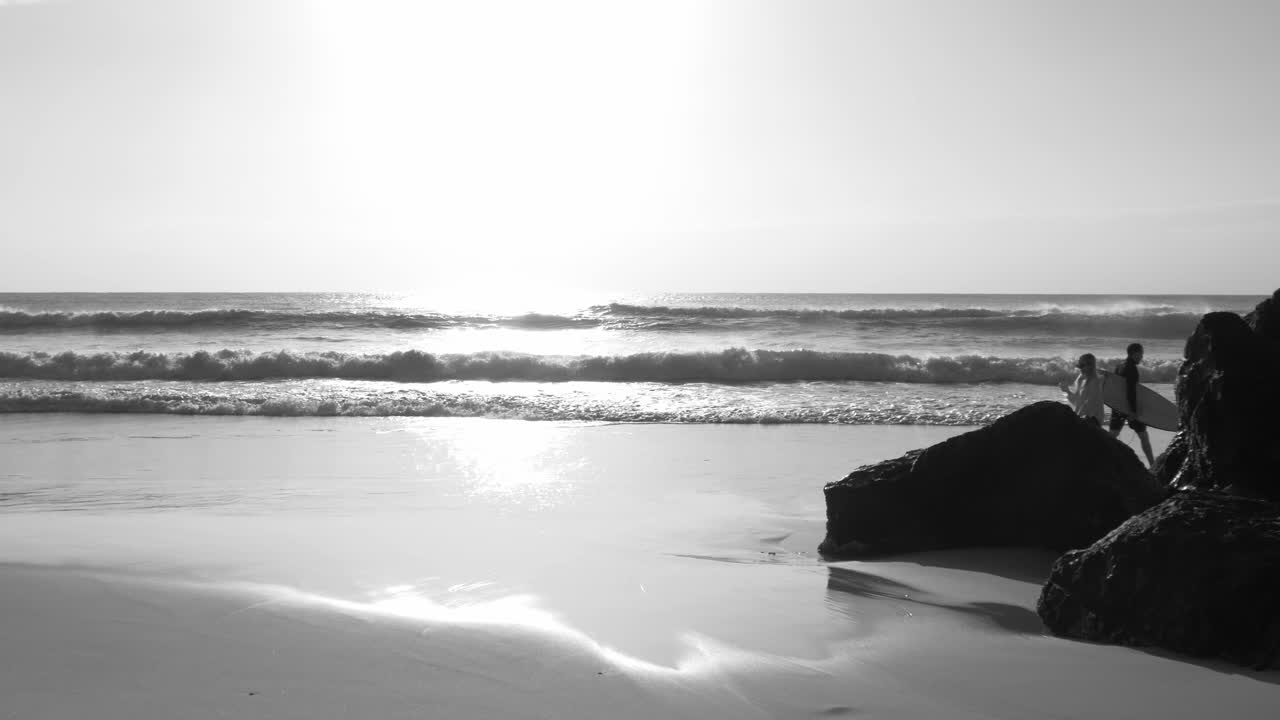 Black And White -  Surfer With Surfboard Walking On The Beach Shore With Bright Sky And Ocean Waves In The Background - Palm Beach In Gold Coast, QLD, Australia - wide slowmo shot